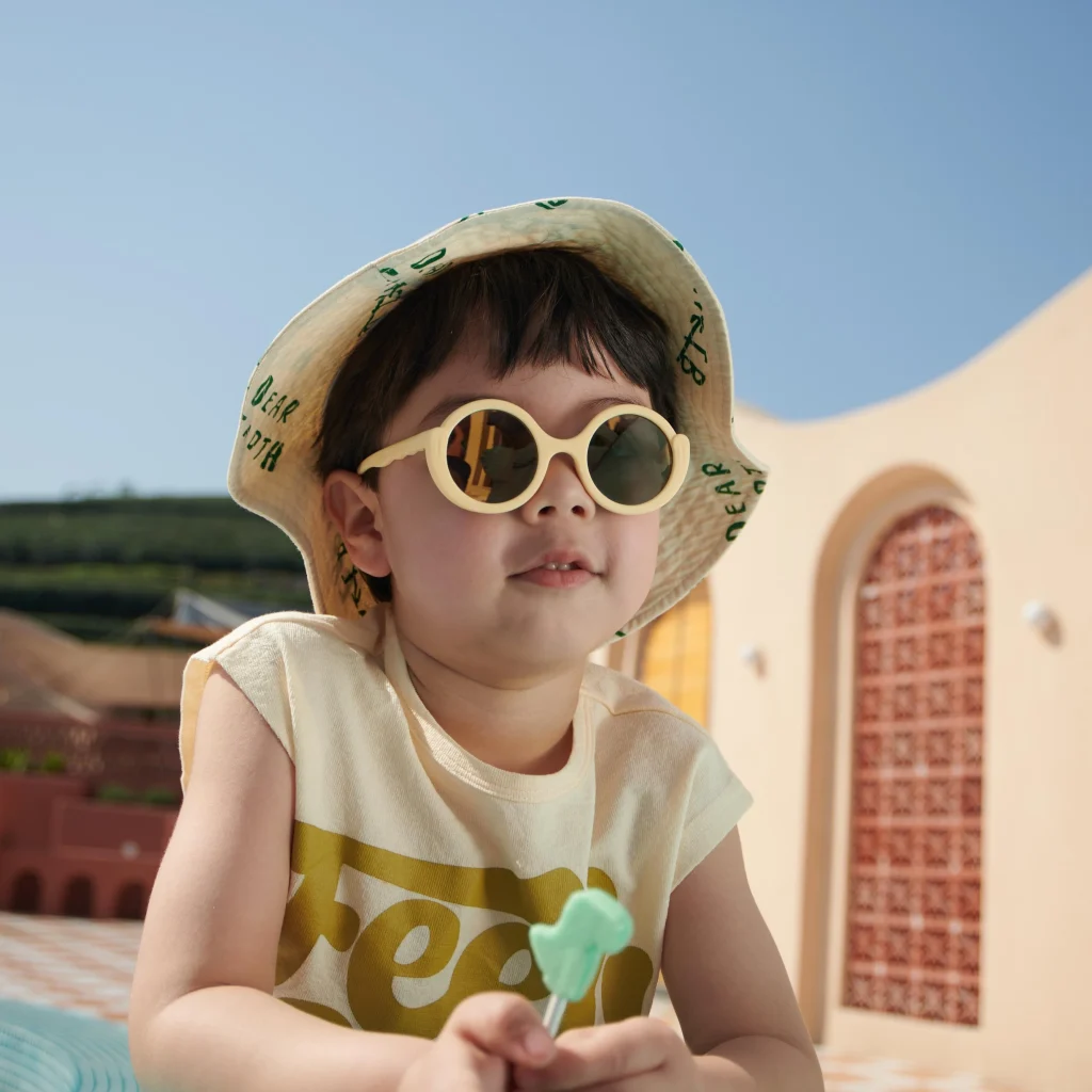 A 4-year-old boy wearing KiGO sunglasses is lying on a swimming ring, holding a lollipop in his hand.