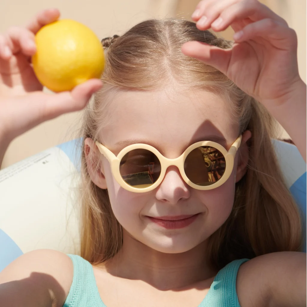 A 5-year-old girl wearing KiGO sunglasses is sitting in a swimming ring, holding a lemon in her hand.