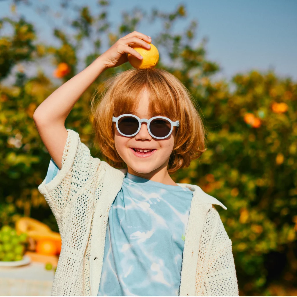A six-year-old boy wearing blue KiGO sunglasses was in an orchard. He held an orange on his head and smiled, showing his teeth.