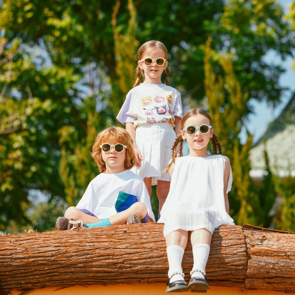 Three children wearing KiGO best kids sunglasses 2025 are sitting on a large, thick log.
