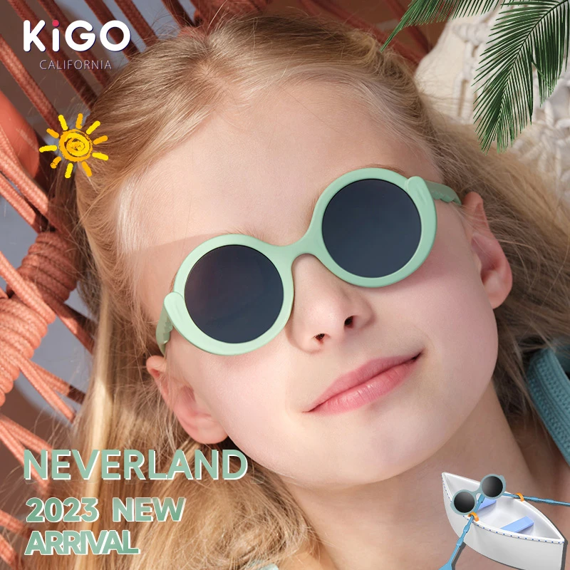 A girl wearing light green KIGO California sunglass end of year kids gifts , with the brand logo in the top left. Text reads "NEVERLAND 2023 NEW ARRIVAL", and there are graphics of a sun and a small boat with sunglasses, set against a summery background with palm leaves.