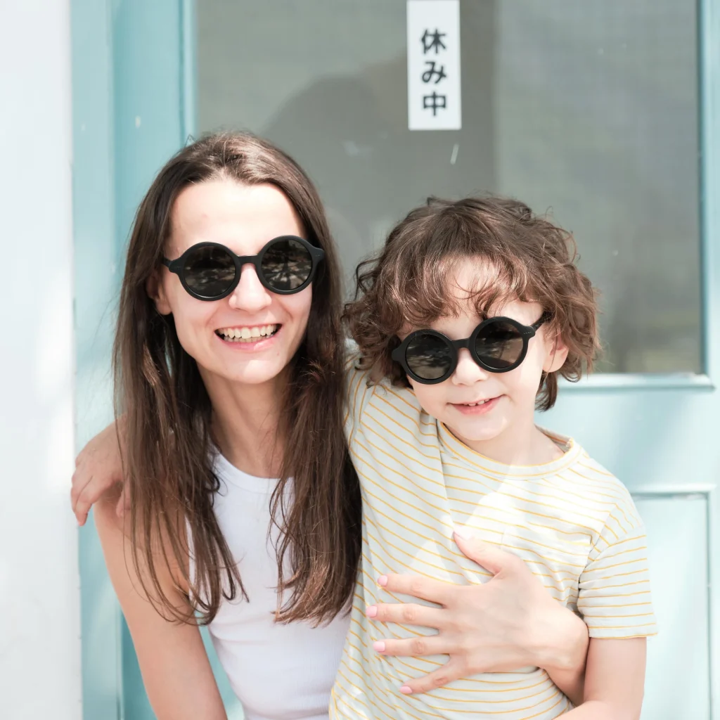 A mother and son are wearing matching KiGO sunglasses in the same color; the mother is holding her son.