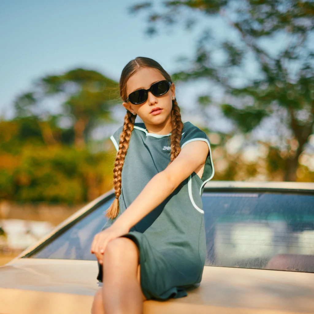 A nine-year-old girl wearing KiGO sunglasses is sitting on the trunk of a car.