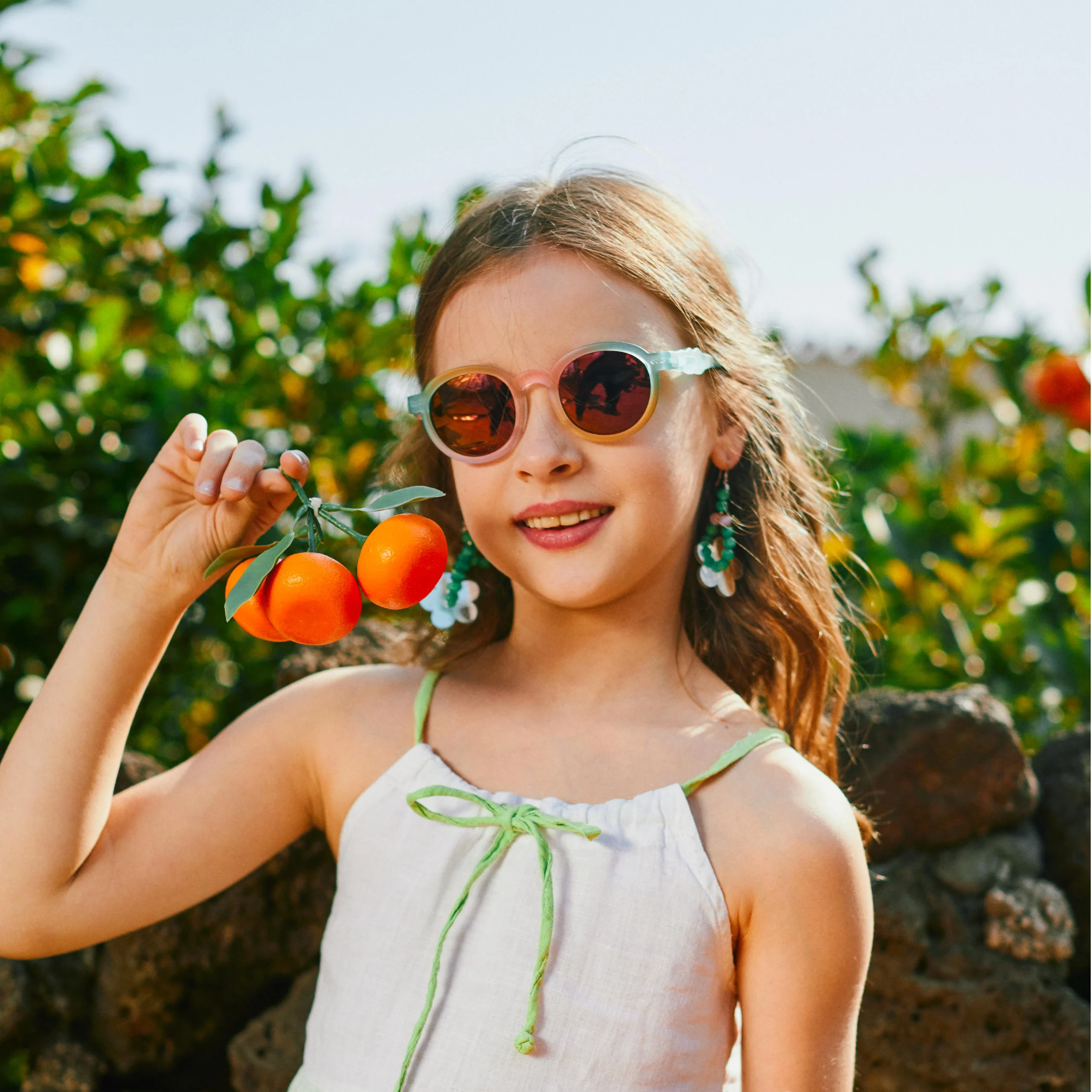 A little girl wearing KiGO kids designer sunglasses is holding three oranges.