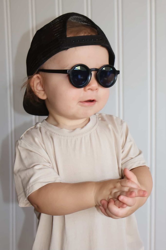 Toddler wearing black toddler sunglasses and a backward black cap, standing in front of a bright white background, smiling happily.