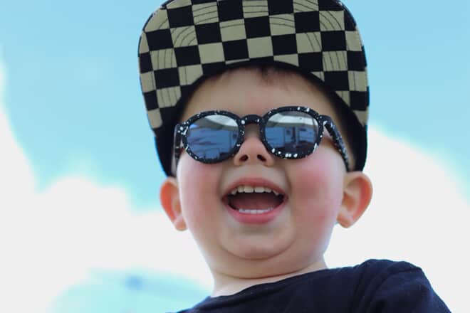 Smiling toddler wearing black sunglasses for kids and a checkered cap, enjoying a sunny day under a bright blue sky.