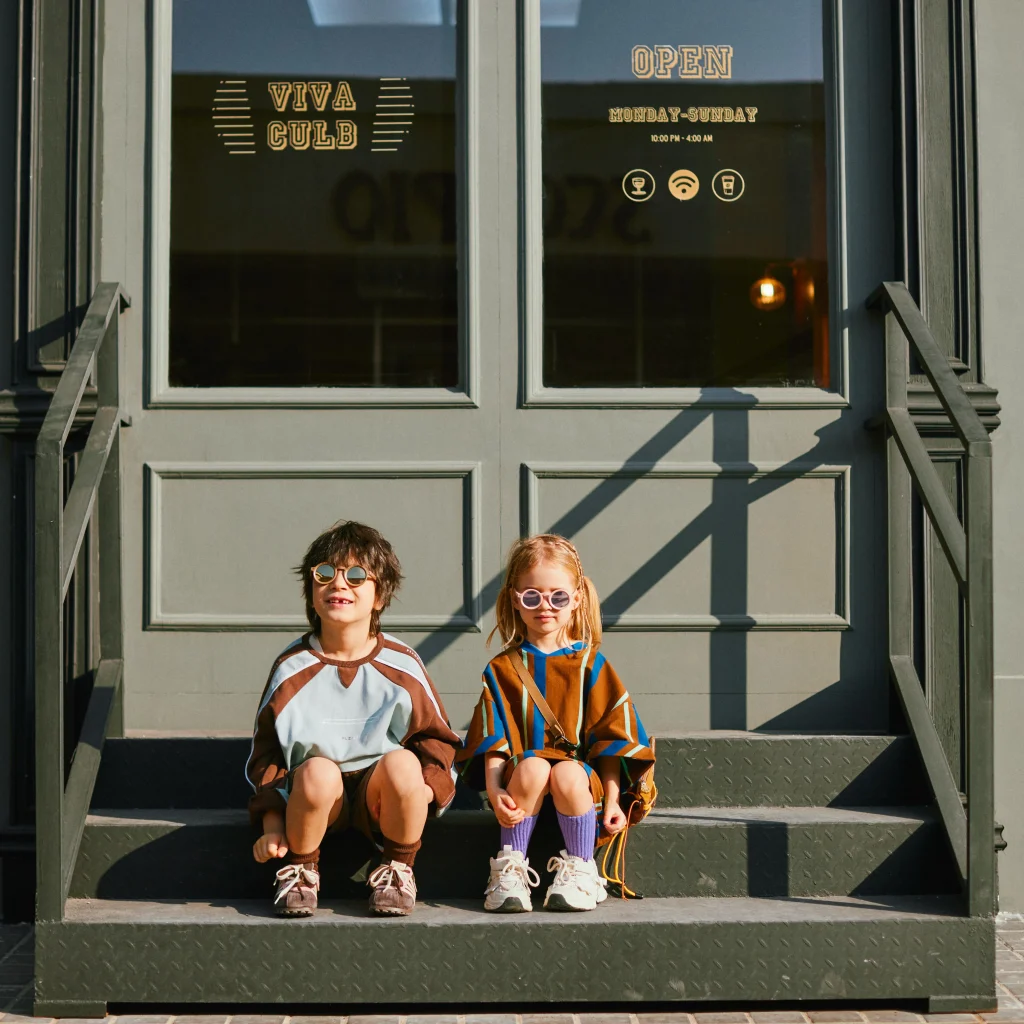Two children wearing KiGO best sunglasses for kids active lifestyles 2025 are sitting on the doorstep.