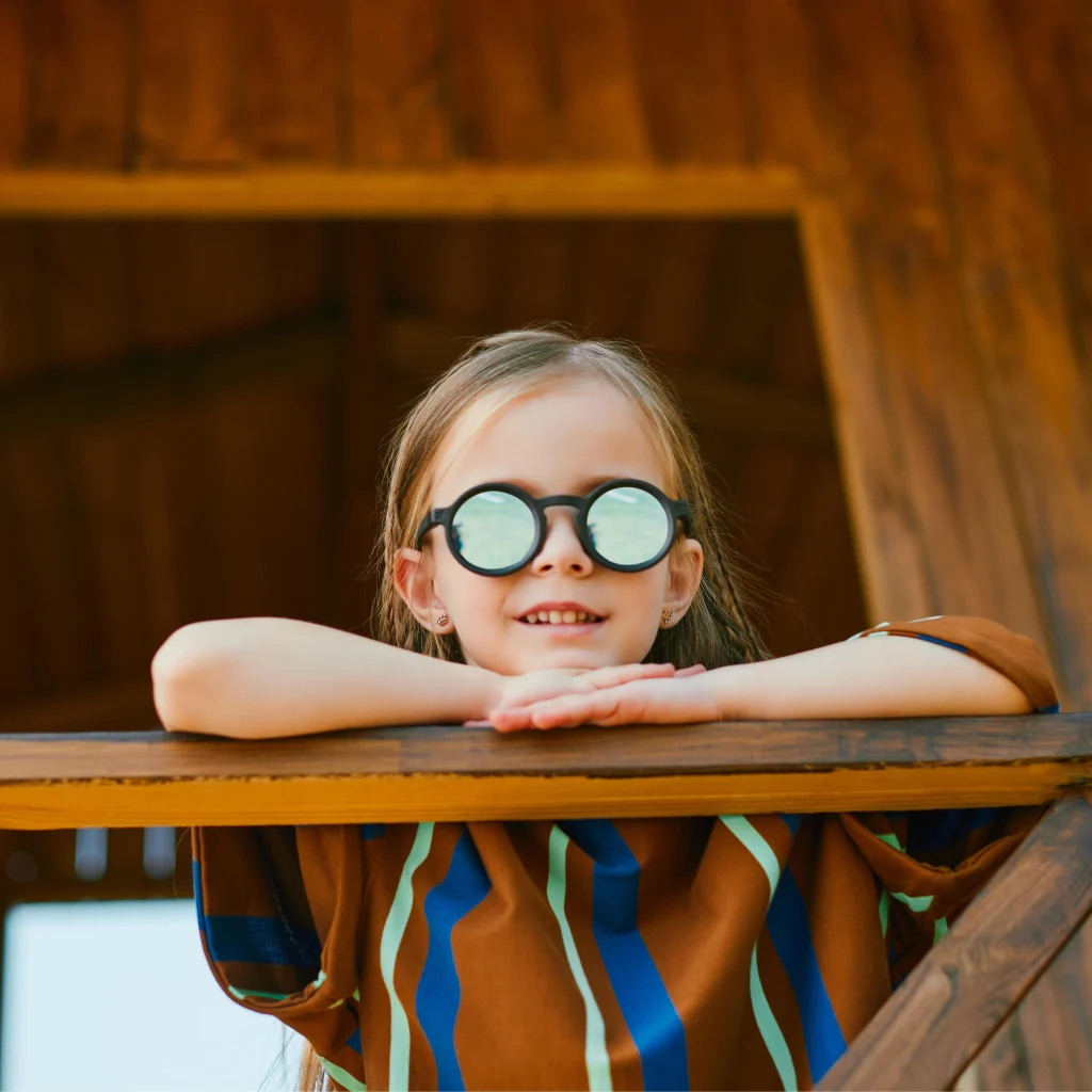A four-year-old girl wearing black KiGO California bendable kids sunglasses​ is leaning on a wooden railing, looking down slightly.