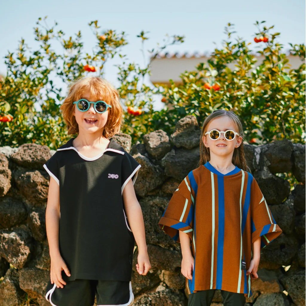 A 2-5 year old boy and a girl wearing green and white KiGO California bendable sunglasses for kids stand side by side in front of a stone wall, smiling and looking ahead.