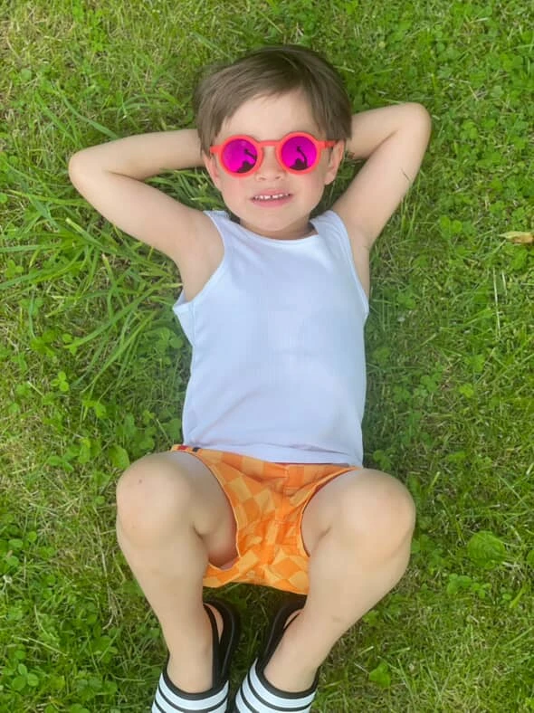 A little boy wearing KiGO California bendable kids sunglasses lies on the grass and looks at the sky.