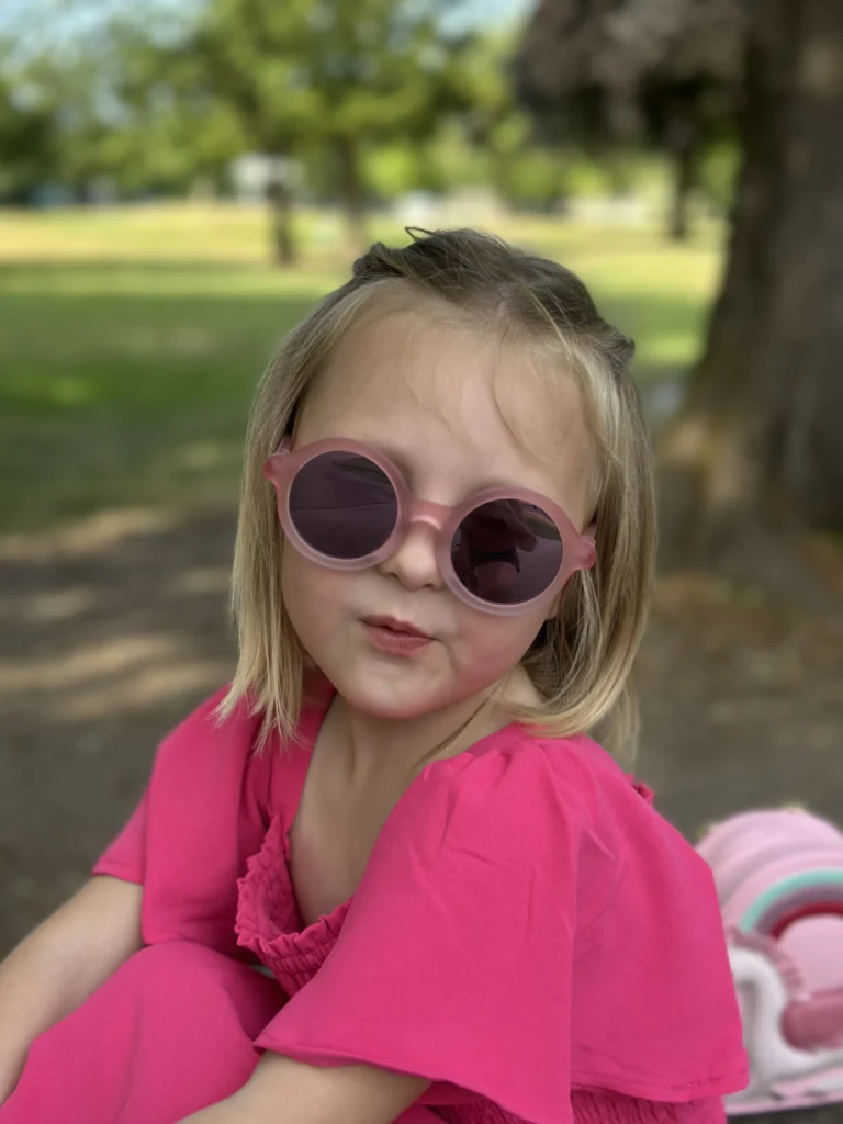 A cute little girl wearing KiGO California 6-9 years old kid blenders sunglasses sitting under a tree.