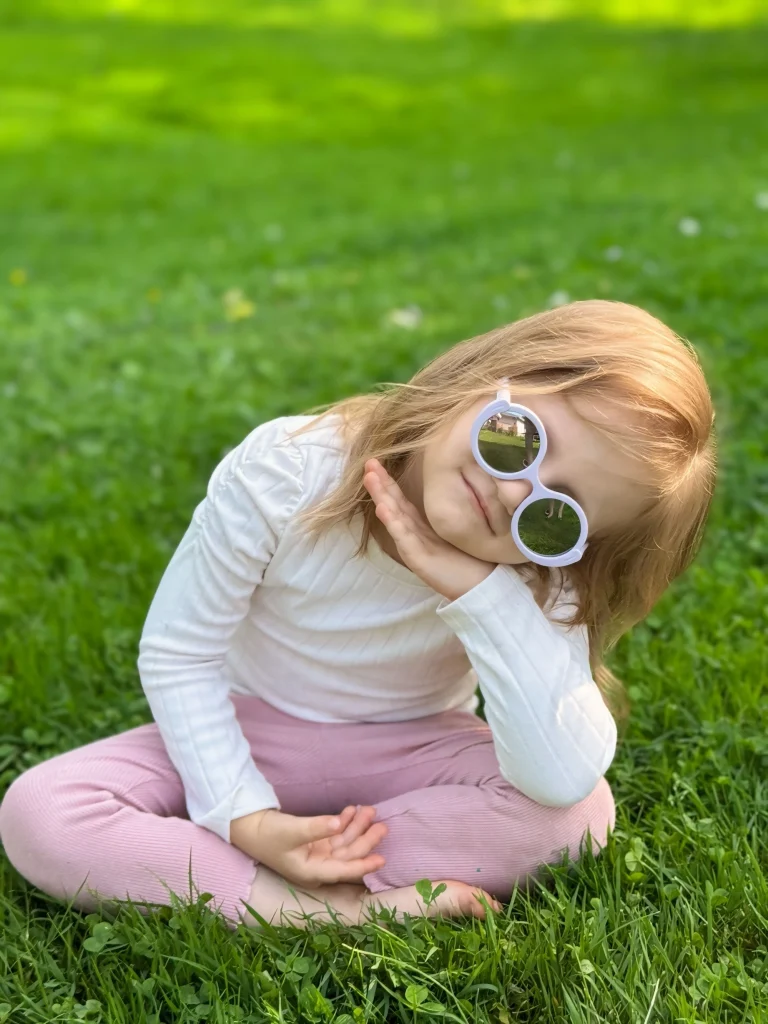 A pretty girl wearing KiGO California sunglasses for girls kids sits on the grass with her head tilted and smiles sweetly.
