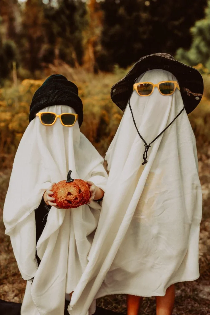 Two children dressed as ghosts with white sheets, wearing yellow sunglasses—one with a black beanie holding a glittery pumpkin, the other with a black bucket hat. They stand in an autumnal, grassy outdoor setting, creating a playful Halloween scene.