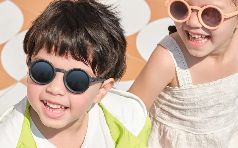 A boy and a girl two babies wearing KiGO baby sunglasses smile under the sun