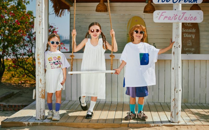 Three children wearing stylish uva uvb sunglasses kids stand and play near a beach - style hut. One child is on a rope swing, and there are signs for "Ice Cream", "Fruit Juice", and "Seaside Diary". The scene is bright and sunny, with greenery and flowers in the background, creating a cheerful, summery vibe.
